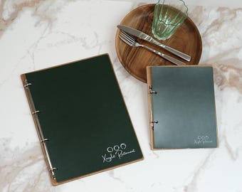 May include: Two dark green menu covers with wooden trim and metal ring binders. The larger menu cover has the text "Kayla's Restaurant" in white script. A wooden plate with silverware and a glass are also present.