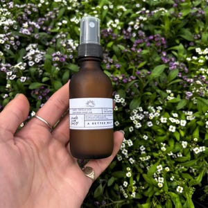 May include: A brown glass spray bottle with a white label is held in a hand. The label reads "Lemon + Vanilla Bean, Custom Spray 002" and "A Better Way". The background is a field of small white and purple flowers.