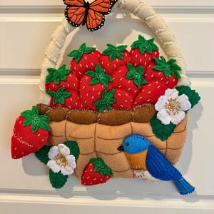 Puede incluir: Decoraci&oacute;n de cesta de fresas de fieltro con asa color crema, llena de fresas rojas y hojas verdes. Se adjuntan una mariposa monarca y un p&aacute;jaro azul. La palabra "Welcome" est&aacute; en una fresa.
