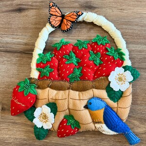 Puede incluir: Una cesta de fresas de fieltro con un asa blanca, llena de fresas rojas y hojas verdes. Una mariposa se posa en el asa, y un mirlo azul y flores blancas decoran la cesta. Una fresa tiene la palabra "Welcome".