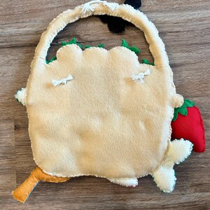 Puede incluir: Una bolsa de fieltro beige con asa, bordes festoneados verdes, una fresa roja y un peque&ntilde;o detalle naranja y azul. La bolsa tiene detalles de borlas blancas y un acento negro.