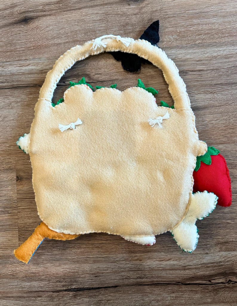 Puede incluir: Una bolsa de fieltro beige con asa, bordes festoneados verdes, una fresa roja y un peque&ntilde;o detalle naranja y azul. La bolsa tiene detalles de borlas blancas y un acento negro.