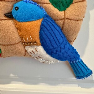 Puede incluir: Un p&aacute;jaro azul de fieltro con pecho naranja y vientre blanco, posado en un nido de fieltro. El p&aacute;jaro tiene un pico negro y est&aacute; adornado con cuentas. El nido es marr&oacute;n y tiene una hoja con lentejuelas blancas.