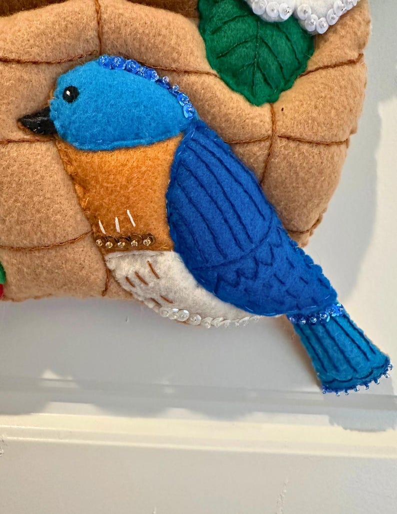 Puede incluir: Un p&aacute;jaro azul de fieltro con pecho naranja y vientre blanco, posado en un nido de fieltro. El p&aacute;jaro tiene un pico negro y est&aacute; adornado con cuentas. El nido es marr&oacute;n y tiene una hoja con lentejuelas blancas.