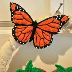 Puede incluir: Una mariposa de fieltro con alas naranjas y contornos negros, acentuada con puntos blancos. La mariposa forma parte de una corona decorativa, con una fresa roja y hojas verdes debajo.