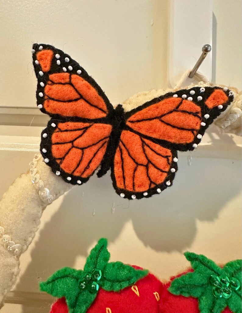 Puede incluir: Una mariposa de fieltro con alas naranjas y contornos negros, acentuada con puntos blancos. La mariposa forma parte de una corona decorativa, con una fresa roja y hojas verdes debajo.