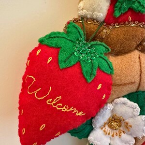 Puede incluir: Una corona de fresas de fieltro con una fresa roja bordada con la palabra "Welcome" en amarillo. La corona presenta hojas verdes, flores blancas y detalles en fieltro marr&oacute;n. Adornada con lentejuelas y cuentas.