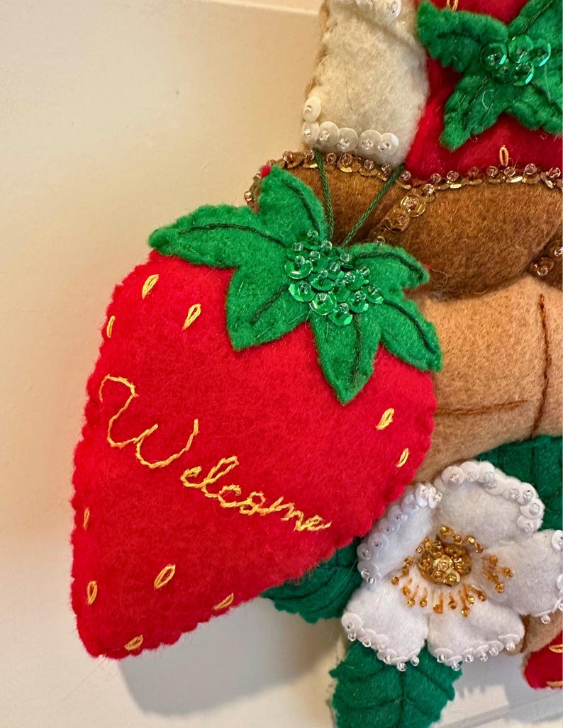 Puede incluir: Una corona de fresas de fieltro con una fresa roja bordada con la palabra "Welcome" en amarillo. La corona presenta hojas verdes, flores blancas y detalles en fieltro marr&oacute;n. Adornada con lentejuelas y cuentas.