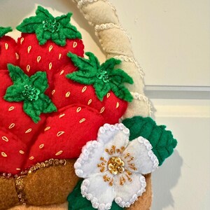 Puede incluir: Una cesta de fresas de fieltro con fresas rojas, hojas verdes y una flor blanca con detalles dorados. La cesta tiene un asa beige y est&aacute; decorada con cuentas doradas. Las fresas tienen peque&ntilde;os detalles de semillas amarillas.