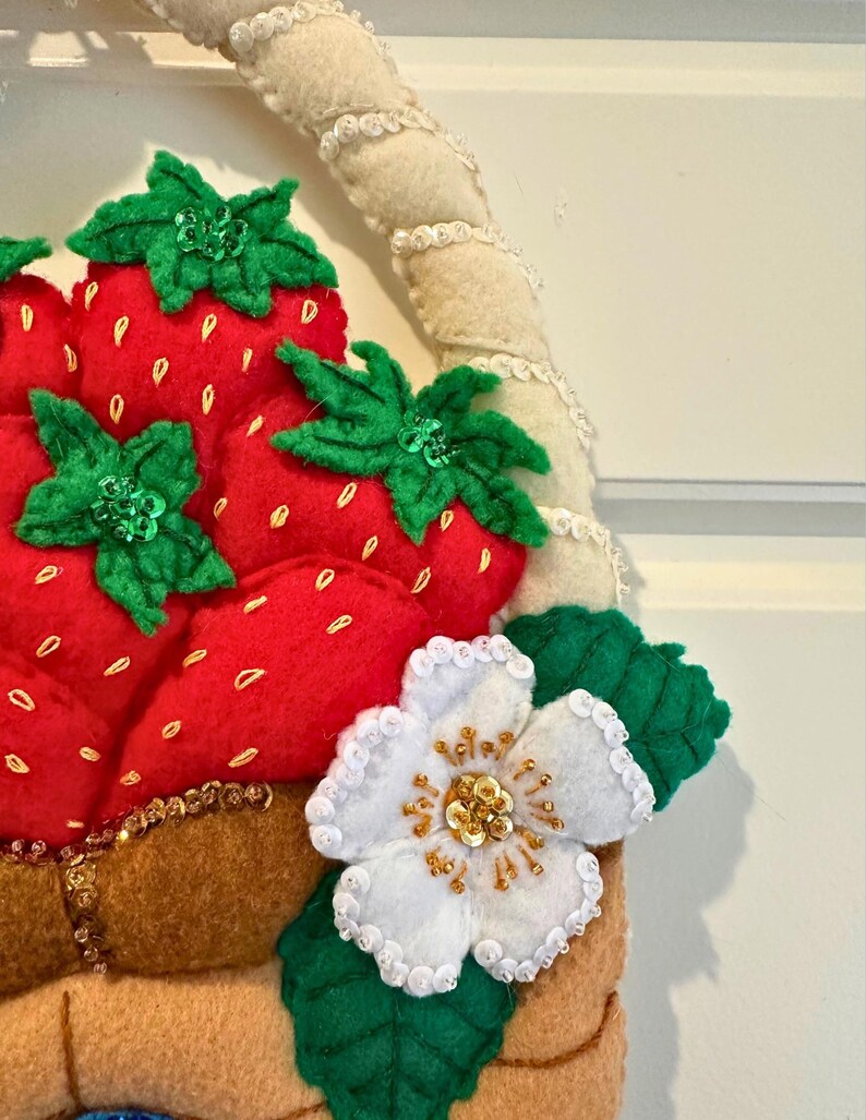 Puede incluir: Una cesta de fresas de fieltro con fresas rojas, hojas verdes y una flor blanca con detalles dorados. La cesta tiene un asa beige y est&aacute; decorada con cuentas doradas. Las fresas tienen peque&ntilde;os detalles de semillas amarillas.
