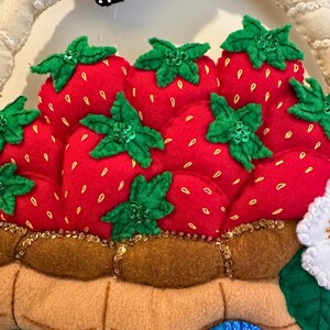 Puede incluir: Una cesta de fresas de fieltro con fresas rojas, hojas verdes y lentejuelas doradas. Tambi&eacute;n se ven una mariposa y un p&aacute;jaro azul. La cesta es de color marr&oacute;n claro y tiene un asa blanca.
