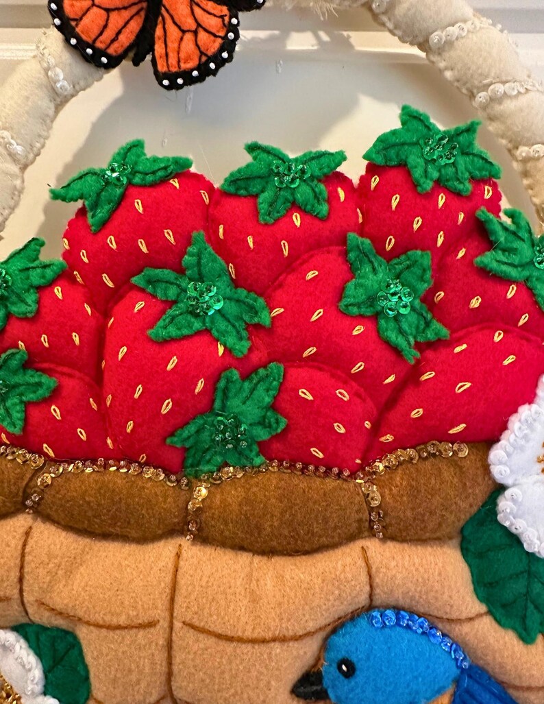 Puede incluir: Una cesta de fresas de fieltro con fresas rojas, hojas verdes y lentejuelas doradas. Tambi&eacute;n se ven una mariposa y un p&aacute;jaro azul. La cesta es de color marr&oacute;n claro y tiene un asa blanca.