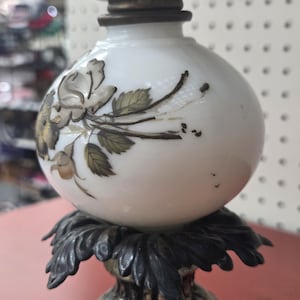 May include: A white glass oil lamp base featuring a hand-painted floral design with brown and grey roses and leaves. The lamp has a dark metallic base shaped like a cluster of leaves and a silver-toned metal collar at the top of the glass globe.