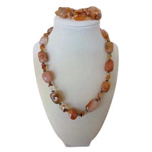 May include: A necklace and bracelet set featuring large, polished, orange-brown gemstone beads. The necklace and bracelet are displayed on a white jewelry bust. The beads are strung together with small, silver-colored connectors.