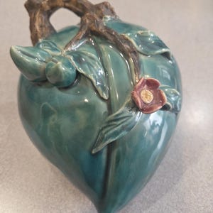 May include: A teal ceramic decorative piece shaped like a fruit, possibly a peach, with a brown branch handle. The piece features green leaves and a small pink flower with a yellow center. The ceramic has a glossy finish.