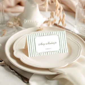 May include: A folded white place card featuring a sage green vertical striped pattern. The center displays a decorative frame with the printed name Mary Mackenzie and the text TABLE 4 in a simple black serif font, centered on a white plate.