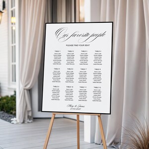 May include: A framed seating chart on a wooden easel. The white sign has black text that reads "Our favorite people" and "Please find your seat." Names are listed under table numbers. The date "23.02.2028" is at the bottom.