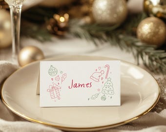 Puede incluir: Una tarjeta de sitio blanca plegada con el nombre James en letra roja, rodeado de ilustraciones de línea en rojo y verde de un regalo, adornos, un gorro de Papá Noel, un bastón de caramelo, un árbol de Navidad y corazones en un plato crema.