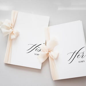 May include: Two white vow books, one labeled "His" and the other "Her", with elegant script. Each book is tied with a cream-colored ribbon and bow. A soft, romantic aesthetic for a wedding.