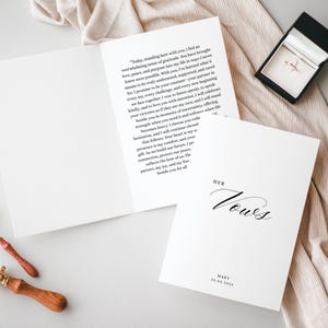 Può includere: Un libretto per i voti nuziali bianco con il testo HER VOWS, MARY e 23.03.2023 stampato sulla copertina. Il libretto aperto mostra un voto scritto sincero. Un portagioie nero contenente un anello d'oro si trova vicino, con un sigillo in legno.
