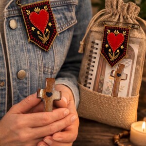 May include: A wooden cross with a black heart detail is held in front of a denim jacket. A banner pin with a red heart and floral design is pinned to the jacket. A burlap bag with a similar banner pin and a notebook is also visible.