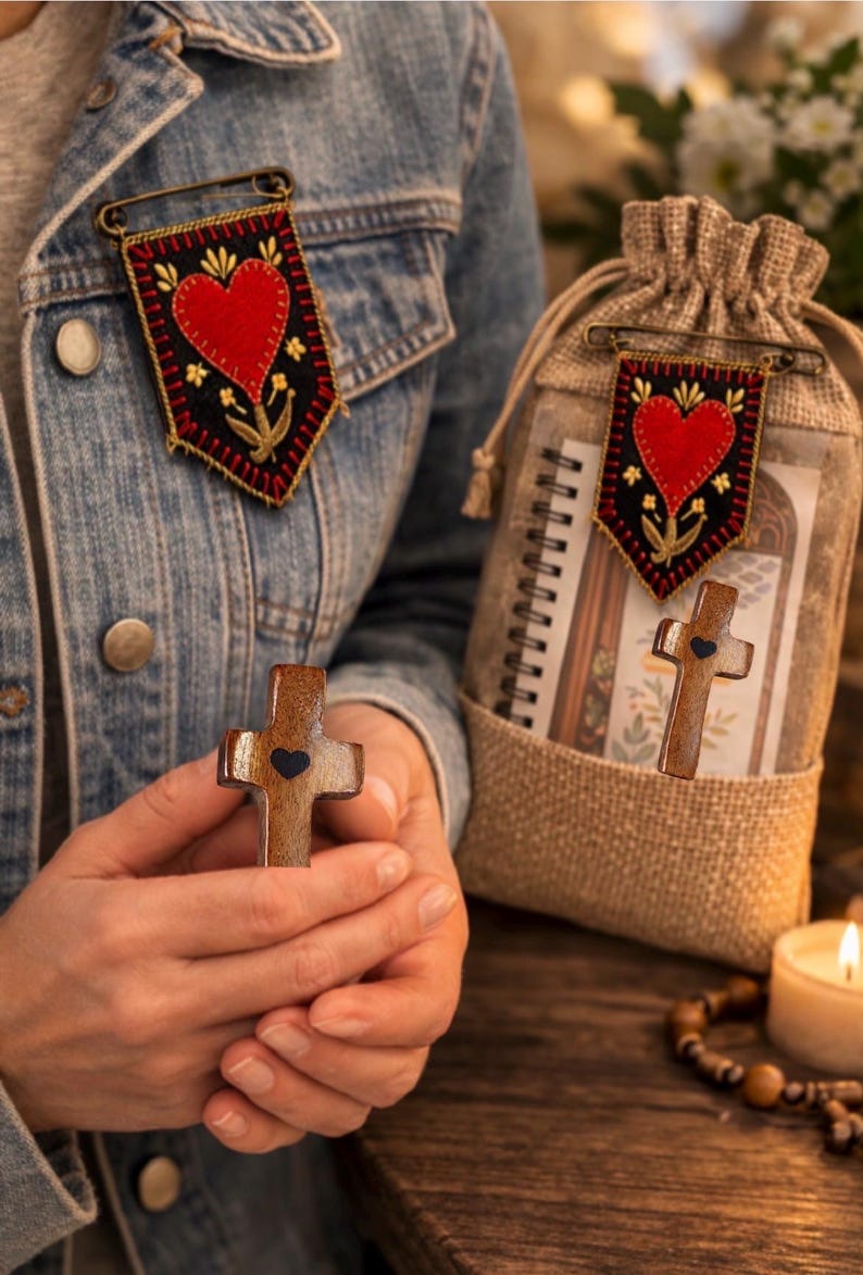 May include: A wooden cross with a black heart detail is held in front of a denim jacket. A banner pin with a red heart and floral design is pinned to the jacket. A burlap bag with a similar banner pin and a notebook is also visible.