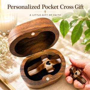 May include: A dark brown wooden oval box with a hinged lid containing a smooth wooden cross with a small black heart inlay. A matching wooden cross with a black heart inlay is held nearby. The text reads Personalized Pocket Cross Gift.
