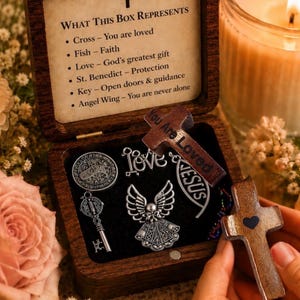 May include: A wooden box with a cross on the lid, open to reveal religious charms and a wooden cross. The box contains a key, angel wing, and other charms. Text on the lid reads "WHAT THIS BOX REPRESENTS".
