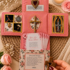 May include: A pink fabric devotional altar featuring a central crucifix, a sacred heart emblem, and religious imagery. It includes cards with text from Joshua 1:9, Deuteronomy 31:8, and The Lord's Prayer. A small wooden cross is held in the foreground.
