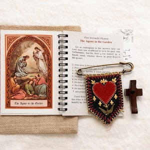 May include: A spiral-bound booklet with the title "The Agony in the Garden" next to a decorative banner with a red heart and a small wooden cross. A safety pin is attached to the banner.