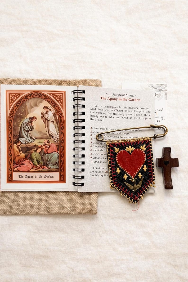 May include: A spiral-bound booklet with the title "The Agony in the Garden" next to a decorative banner with a red heart and a small wooden cross. A safety pin is attached to the banner.
