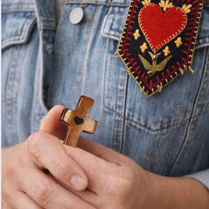 May include: A wooden cross with a heart-shaped cutout is held in the hands. A decorative pin with a red heart and gold accents is pinned to a denim jacket. The cross is brown, and the pin has a black background.