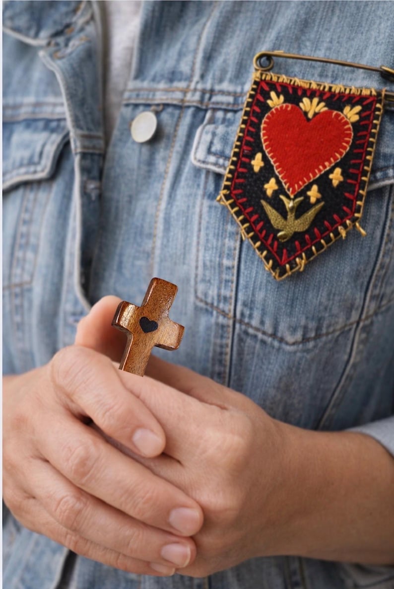 May include: A wooden cross with a heart-shaped cutout is held in the hands. A decorative pin with a red heart and gold accents is pinned to a denim jacket. The cross is brown, and the pin has a black background.