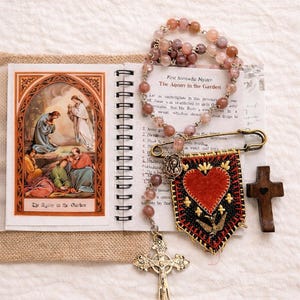 May include: An open spiral-bound book with an illustration of a religious scene, a rosary, a banner with a heart, and a wooden cross. The rosary has pink beads. The banner has a red heart and a gold cross. The text "The Agony in the Garden" is visible.