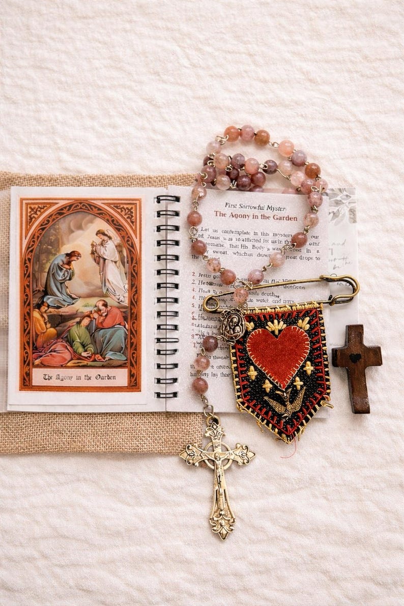May include: An open spiral-bound book with an illustration of a religious scene, a rosary, a banner with a heart, and a wooden cross. The rosary has pink beads. The banner has a red heart and a gold cross. The text "The Agony in the Garden" is visible.