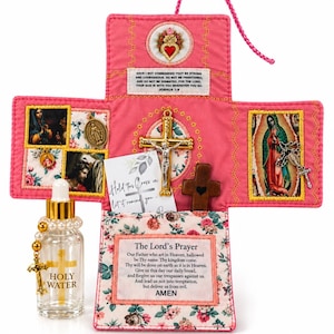 May include: A pink fabric cross-shaped prayer kit featuring religious imagery, a gold crucifix, and a wooden cross. It includes a clear glass bottle labeled Holy Water with a gold dropper and a rosary. Text includes Joshua 1:9, The Lord's Prayer, and Hold This Cross in your hand.
