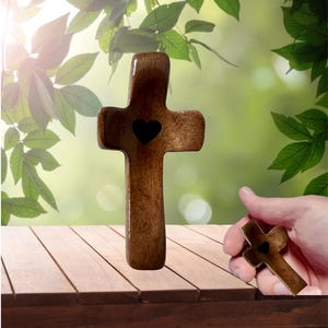 May include: A wooden cross with a heart-shaped cutout in the center. The cross is brown and has a smooth finish. A hand holds a smaller version of the cross. The background features green foliage and a wooden surface.