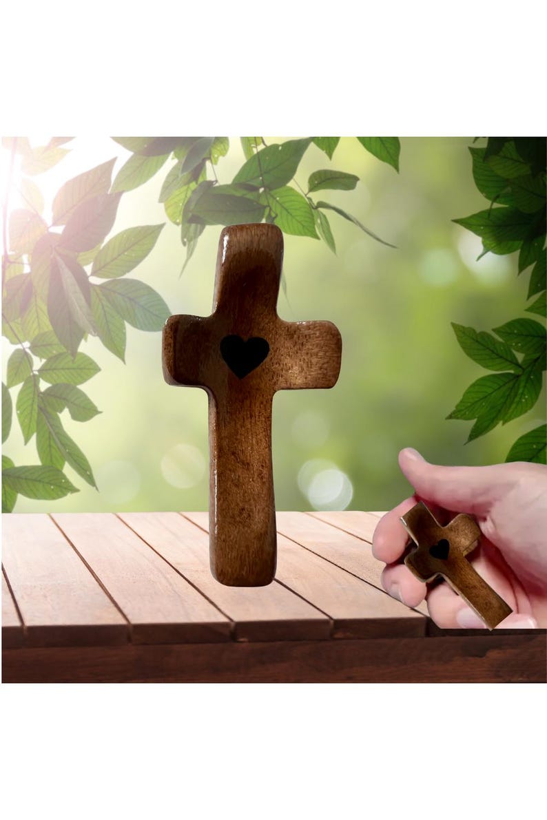 May include: A wooden cross with a heart-shaped cutout in the center. The cross is brown and has a smooth finish. A hand holds a smaller version of the cross. The background features green foliage and a wooden surface.