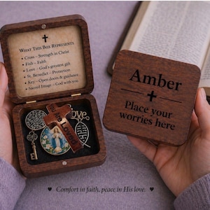 May include: A wooden keepsake box with a hinged lid, open to reveal religious charms and a cross. The lid of the box has text that reads "WHAT THIS BOX REPRESENTS". A separate wooden square with the name "Amber" and the text "Place your worries here" is held nearby.