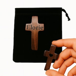 May include: Two wooden cross pendants, one with the name "Elorie" engraved, and the other with a heart cutout. The crosses are dark brown and displayed against a black pouch. One cross is held in a hand.