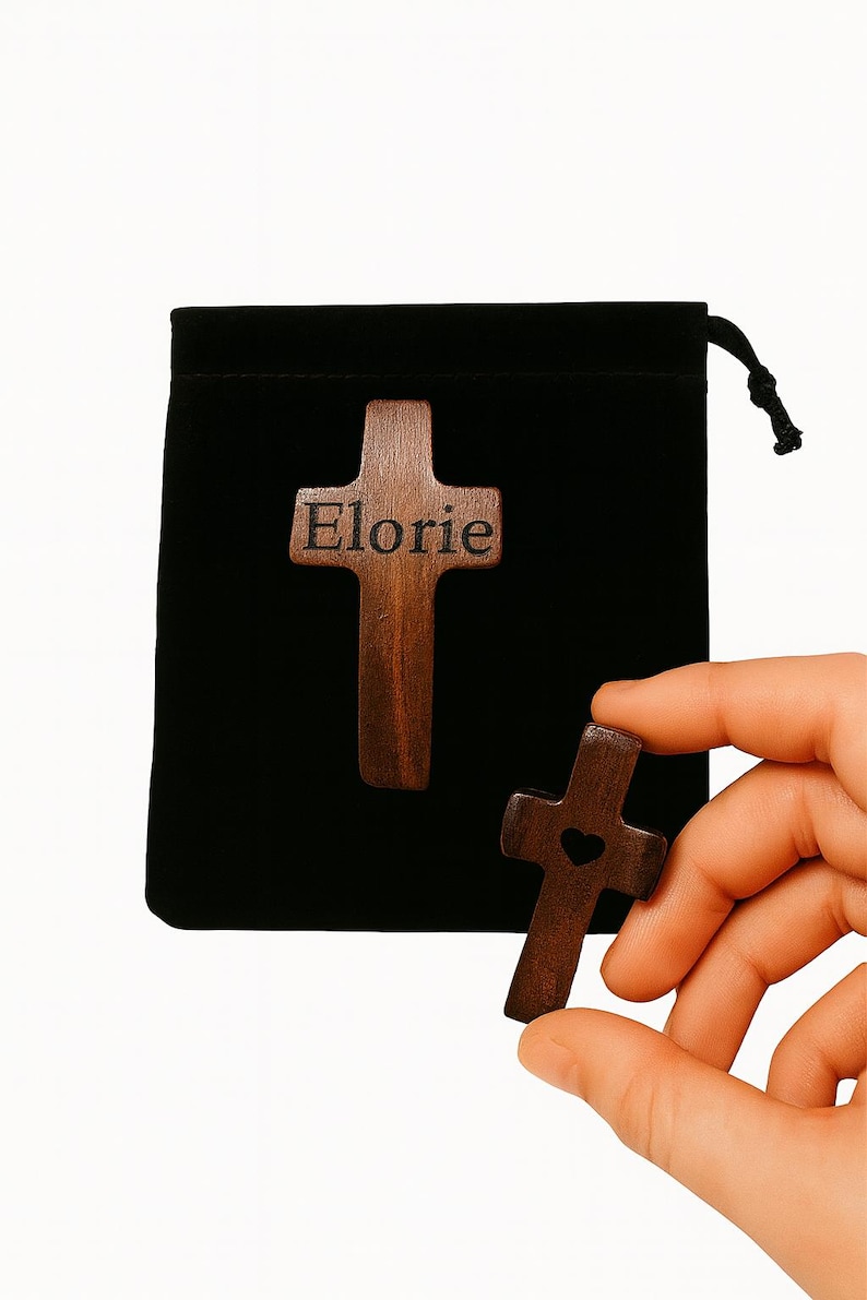 May include: Two wooden cross pendants, one with the name "Elorie" engraved, and the other with a heart cutout. The crosses are dark brown and displayed against a black pouch. One cross is held in a hand.