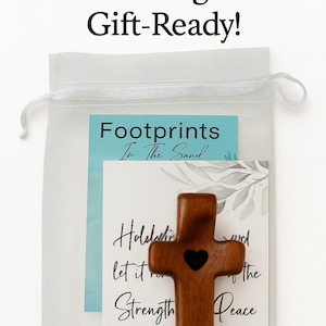 May include: A gift-ready mesh bag containing a wooden cross with a heart cutout, and two cards. The cards have the text "Footprints In The Sand" and a scripture about strength and peace. The bag is white.