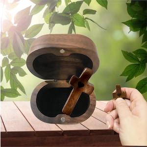 May include: A wooden cross with a heart-shaped cutout rests inside an open, oval-shaped wooden box. A smaller cross is held nearby. The box and crosses are made of a dark wood, and the background is a blurred view of green foliage.