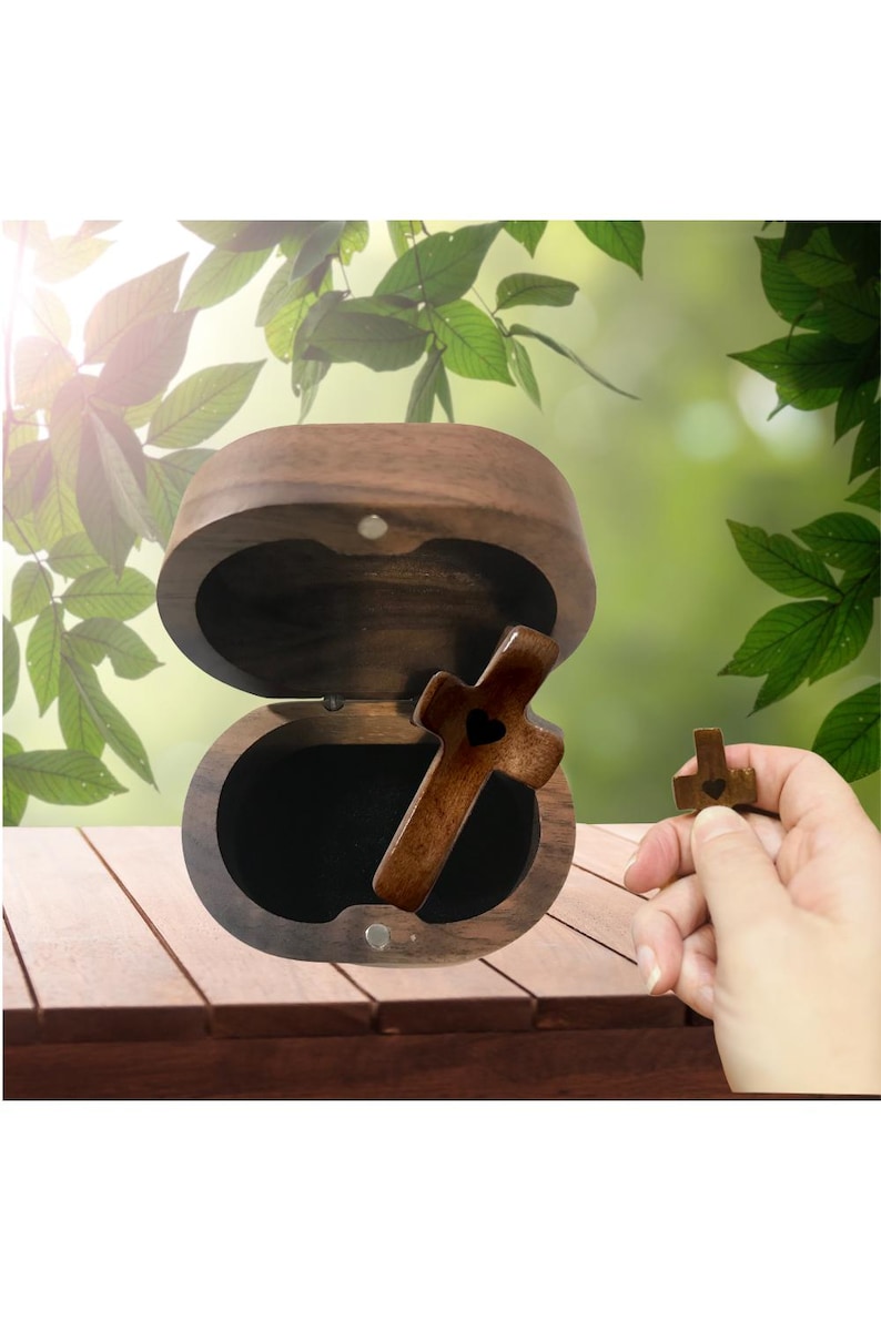 May include: A wooden cross with a heart-shaped cutout rests inside an open, oval-shaped wooden box. A smaller cross is held nearby. The box and crosses are made of a dark wood, and the background is a blurred view of green foliage.