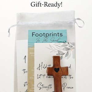May include: A wooden cross with a heart cutout, included with scripture cards and a mesh bag. The cards have text that reads "Footprints in the Sand", "Hold and Let it remind you of the Strength and Peace". The bag is white.