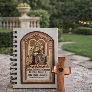 May include: A wooden cross with a black heart and a spiral-bound notebook with religious artwork and text. The notebook cover features an illustration of figures in a church setting. The text reads "The Fifteen Mysteries of The Holy Rosary".