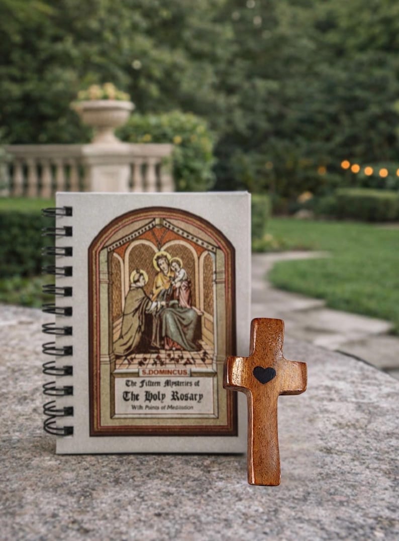 May include: A wooden cross with a black heart and a spiral-bound notebook with religious artwork and text. The notebook cover features an illustration of figures in a church setting. The text reads "The Fifteen Mysteries of The Holy Rosary".