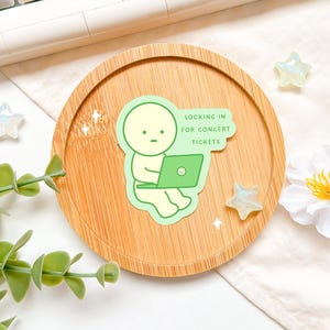 Peut inclure: Un autocollant vert clair représentant un personnage de dessin animé avec un ordinateur portable. Le personnage est assis et a une expression neutre. Le texte sur l'autocollant dit "LOCKING IN FOR CONCERT TICKETS". L'autocollant est sur un plateau en bois avec des étoiles décoratives et une fleur.