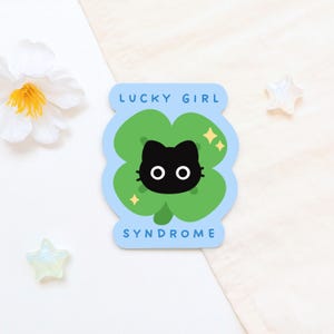 Puede incluir: Una pegatina con la cara de un gato negro dentro de un trébol verde de cuatro hojas. La pegatina tiene un borde azul claro con las palabras "LUCKY GIRL" y "SYNDROME". También se incluyen acentos de estrellas doradas.
