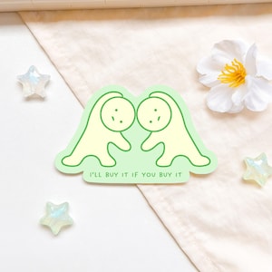 Puede incluir: Una pegatina con dos figuras de dibujos animados de color amarillo claro, delineadas en verde, sobre un fondo verde claro. El texto "I'LL BUY IT IF YOU BUY IT" está en la parte inferior. La pegatina está sobre una superficie beige con decoraciones en forma de estrella.